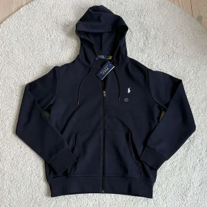 Mörkblå hoodie från Polo Ralph Lauren - Helt ny Snygg mörkblå hoodie från Polo Ralph Lauren med dragkedja, huva med snörning och klassisk broderad logga på bröstet. Tröjan har känguruficka och ribbade muddar. Perfekt för en chill och stilren look.