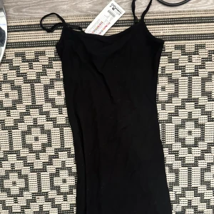 Svart tight långklänning med smala band - En svart, tight långklänning med smala axelband och enkel, stilren design. Perfekt för dig som gillar minimalistisk stil och vill ha en klänning som sitter snyggt och markerar figuren. Gjord i ett mjukt och stretchigt material för extra komfort. Aldrig använd, endast legat i garderoben. Prislapp kvar.