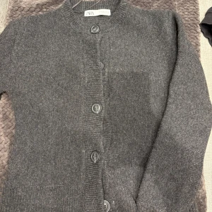 Mörkgrå kofta från Zara i S - Mysig mörkgrå kofta från Zara med rund halsringning och stora knappar framtill. Stickad i mjukt material och har långa ärmar. Perfekt att slänga över en t-shirt eller linne för en chill look.