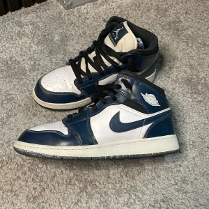 Nike Air Jordan 1 Mid Navy/Vit - Snygga Nike Air Jordan 1 Mid sneakers i marinblått och vitt skinn. Klassisk siluett med svart snörning, vit mellansula och mörk yttersula. Ikonisk vinge-logga på sidan och Nike swoosh. Perfekta för dig som vill sticka ut med en tidlös streetstyle. Storlek 40