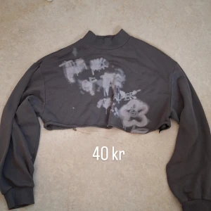 Grå croppad sweatshirt med graffiti - Grå croppad sweatshirt med rå kant och unikt graffiti-inspirerat tryck i vitt och ljusrosa framtill. Tröjan har lång ärm och ribbad mudd vid ärmslut samt högre krage. Perfekt för en edgy streetstyle-look.