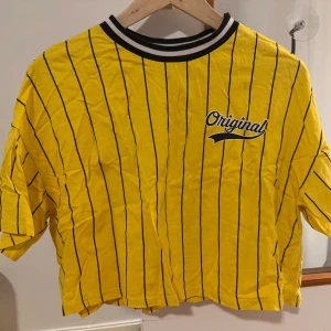 H&M Gul randig croppad t-shirt  - Gul croppad t-shirt med svarta vertikala ränder och trycket 'Original' samt siffran 79 på ryggen. T-shirten har korta ärmar, sportig ribbad krage i svart och vitt och en loose fit. Från H&M i storlek S. 