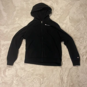 Svart Champion hoodie med dragkedja - Svart hoodie från Champion i storlek Cm174/179  med hel dragkedja och huva. Broderad Champion-logga på bröstet och tryckt logga på ena ärmen. Mjuk insida och klassisk passform, perfekt för chill eller streetwear.