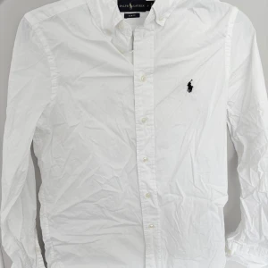 Vit skjorta från Ralph Lauren slim fit - Klassisk vit skjorta från Ralph Lauren i slim fit-modell. Skjortan har lång ärm, knappar framtill och den ikoniska broderade loggan på bröstet. Tillverkad i bomull och passar perfekt till en stilren look.