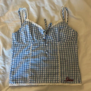 Blå rutig linnetopp från Guess - Supersöt blå och vit rutig linnetopp från Guess med broderad logga. Toppen har smala axelband, knytning framtill, dekorativa knappar och spetsdetaljer längs kanterna. Perfekt för en somrig look med vintagevibbar.