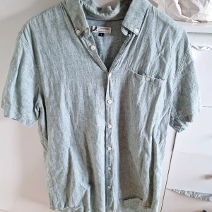 Grön linneskjorta från Dressmann - Snygg grön skjorta i linnemix från Dressmann. Skjortan har korta ärmar, klassisk krage och knäppning framtill med vita knappar. Perfekt för varma dagar och ger en avslappnad vibe. Bröstficka med knappdetalj.