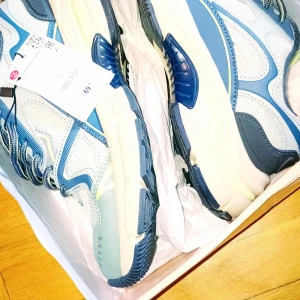 Blå och vita sneakers med meshdetaljer - Snygga sneakers i blått och vitt med coola meshpaneler och detaljer i olika blå nyanser. Skorna har snörning och en chunky, beige sula med blå inslag. Perfekta för dig som gillar sportig stil och vill sticka ut lite extra.