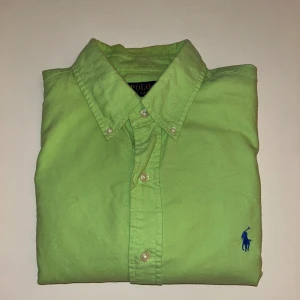Grön skjorta från Polo Ralph Lauren - Fräsch grön skjorta från Polo Ralph Lauren i slim fit. Skjortan har klassisk krage, knäppning framtill och blå broderad logga på bröstet. Tillverkad i mjuk bomull och har långa ärmar. Perfekt för dig som gillar stilrena och färgglada plagg.