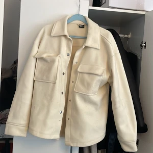 Overshirt i beige från Gina Tricot - Säljer en oversized beige overshirt från Gina Tricot i mjukt material. Jackan har stora bröstfickor, klassisk krage och knappar framtill. Perfekt att slänga över en hoodie eller t-shirt för en chill look.