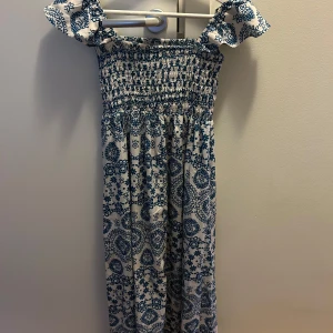 Vit och blå långklänning med mönster - Supersöt långklänning i vitt med blått mönster och smockad överdel. Klänningen har offshoulder-ärmar med volang och är gjord i mjuk bomull. Perfekt för varma dagar när du vill känna dig både chill och snygg. Har även en till i större storlek om det önskas eller om man vill matcha 