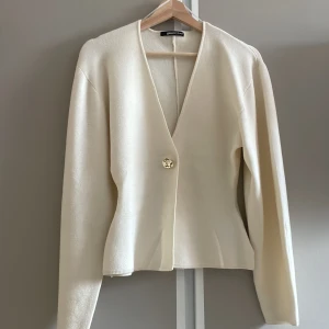 Krämvit kofta från Gina Tricot XS - Snygg krämvit kofta från Gina Tricot i storlek XS. Koftan har en djup v-ringning och stängs med en dekorativ guldig knapp framtill. Långärmad och i mjukt, stickat material som ger en clean och stilren look.