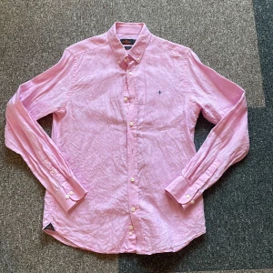 Rosa linneskjorta från Morris - Snygg rosa skjorta från Morris i 100% linne. Klassisk button down-krage, vita knappar och broderad logga på bröstet. Perfekt för dig som gillar en stilren och fräsch look. Skjortan har lång ärm och normal passform. Ny pris 1300kr, Skjorta är i knappast andvänd.