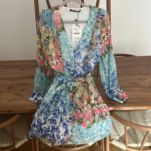 Blommig kortklänning från Zara - Superfin kortklänning från Zara med färgglatt blommönster i blått, rosa, gult och grönt. Klänningen har v-ringning, långa ärmar, knappar framtill och ett matchande knytbälte i midjan. Perfekt för vår och sommar när du vill sticka ut lite extra.