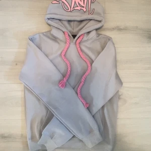 Syna world hoodie - Säljer en grå dyna world hoodie med feta rosa snören och tryck på huvan. Hoodien har en klassisk passform, mjukt material och stor huva. Perfekt för dig som vill sticka ut med streetstyle-vibe och coola detaljer.🥰💯