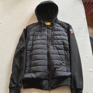 Svart cardigan från Parajumpers - Svart cardigan (Ivor) från Parajumpers med quiltad front och rygg, huva med snörning och dragkedja hela vägen. Ärmar och sidor i mjuk fleece, ribbade muddar och två dragkedjefickor. Märkespatch på ärmen och snygga detaljer vid halsen. Använd 1 gång. Orginal pris 4400kr.