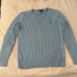 Ljusblå stickad tröja Polo Ralph Lauren - Supersnygg ljusblå stickad tröja från Polo Ralph Lauren med klassiskt kabelstickat mönster och broderad logga på bröstet. Rund halsringning och långa ärmar, perfekt för lager-på-lager-stil under kyliga dagar.
