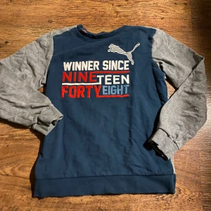 Blå och grå Puma sweatshirt - Snygg sweatshirt från Puma i marinblått med grå ärmar. Framsidan har tryck med texten 'Winner since nineteen forty eight' i vitt, rött och blått samt Puma-logga. Tröjan har rund hals och långa ärmar, perfekt för en sportig look.