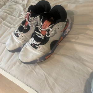 Nike React sneakers i silver och svart - Snygga Nike React sneakers med futuristisk design i silver, svart och orange detaljer. Skorna har vit snörning, rund tå och en unik sula med camo-liknande mönster. Perfekta för dig som gillar streetstyle och vill sticka ut.