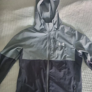 Under Armour grå vindjacka med huva - Snygg tvåfärgad vindjacka från Under Armour i ljusgrått och mörkgrått. Jackan har huva med snörning, dragkedja hela vägen och praktiska fickor. 