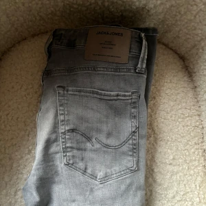 Grå jeans från Jack & Jones - Snygga grå jeans från Jack & Jones med klassisk femficksdesign och diskret bakficka med broderad detalj. Jeansen har en ljusgrå tvätt och är tillverkade i mjukt denimtyg som ger en skön känsla. Perfekta för en avslappnad och stilren look.