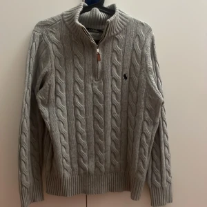 Kabelstickad ralph lauren  - Säljer en grå kabelstickad tröja från Polo Ralph Lauren med half zip och hög krage. Tröjan har lång ärm, ribbade muddar och klassisk logga broderad på bröstet. Perfekt för dig som gillar stilren och tidlös design.