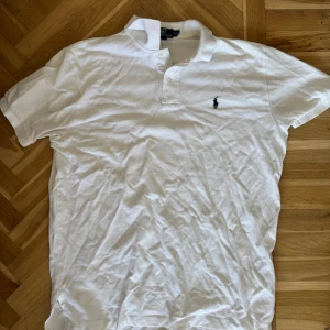 Vit pikétröja från Ralph Lauren - Klassisk vit pikétröja från Ralph Lauren med korta ärmar och den ikoniska broderade loggan på bröstet. Tröjan har krage och knappar framtill, perfekt för en clean och stilren look. Tillverkad i mjuk bomull som andas.