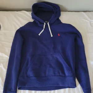 Blå hoodie från Polo Ralph Lauren - Snygg marinblå hoodie från Polo Ralph Lauren med klassisk röd broderad logga på bröstet. Tröjan har huva med vita snören och en stor magficka. Perfekt för dig som gillar stilren och sportig look.