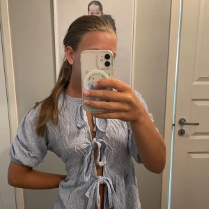 Ljusblå knytblus från Gina Tricot - Supersöt ljusblå blus från Gina Tricot med crinklad struktur och tre knyt framtill. Blusen har korta puffärmar och en luftig känsla, perfekt för varmare dagar. Snygg att styla med jeans eller kjol för en chill look.