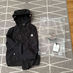 Svart vindjacka från Moncler - Svart vindjacka från Moncler med huva och snörning, halv dragkedja och Moncler-logga på bröstet. Jackan har praktiska fickor med tryckknappar och elastisk nederkant. Perfekt för dig som vill ha en stilren och sportig look.