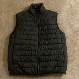 Svart quiltad dunväst med dragkedja - En svart quiltad dunväst med hög krage och hel dragkedja framtill. Västen har en stilren look med horisontella sömmar och är perfekt att slänga över en hoodie eller tröja. Passar dig som gillar enkel och clean stil.