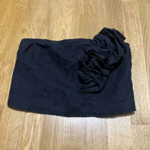 Svart tubtopp med volangdetalj - Snygg svart tubtopp med en stor volangdetalj på ena sidan som ger en cool och trendig look. Toppen är axelbandslös och har en stilren, enkel form som passar perfekt till jeans eller kjol.