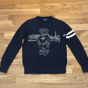 Mörkblå Polo Ralph Lauren Paris-tröja - Mörkblå Polo bear tröja med Paris motiv. Unik design med vitt mot mörkblå bakgrund. Ralph Lauren har gett den en sportig känsla med två vita streck på vänster arm. I perfekt skick, aldrig använd. Pm för fler frågor.