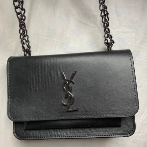 Svart axelväska från Saint Laurent - Snygg svart axelväska från Saint Laurent med klassisk YSL-logga i metall framtill. Väskan har kedjerem i metall och är tillverkad i slätt skinn. Perfekt storlek för det viktigaste och ger en lyxig touch till din outfit. 