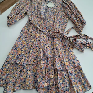 Blommig kortklänning från Valerie - Supersöt kortklänning från Valerie med blommigt mönster i pastelliga toner av blått, rosa och gult. Klänningen har omlottmodell, volangdetaljer, knytband i midjan och trekvartsärm. Tillverkad i en mjuk mix av viskos och cupro – perfekt för vår och sommar.