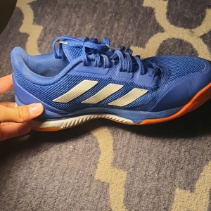 Blå adidas sneakers med orange sula - Snygga blå adidas sneakers med klassiska vita ränder på sidan och orange yttersula. Ovandelen är i mesh och syntet för bra andningsförmåga. Skon har snörning och adidas-logga på plösen. Perfekt för dig som gillar sportig stil.