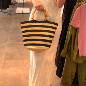 Randig shoppingväska/ strandväska - Snygg shoppingväska med breda svarta och beige ränder och coola handtag i bambu. Väskan har en korgliknande form och är perfekt för dig som vill sticka ut med en trendig accessoar. Passar till alla dina outfits och rymmer allt du behöver.