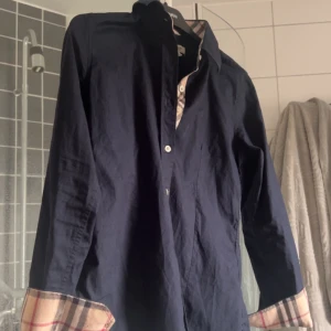 Mörkblå Burberry skjorta med rutiga detaljer - Snygg mörkblå skjorta från Burberry med klassiska rutiga detaljer på insidan av kragen och ärmsluten. Skjortan har lång ärm, knappar framtill och är tillverkad i bomull. Perfekt för dig som gillar stilrena plagg med en twist.storlek S