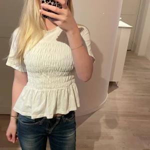 Vit smockad topp från Vero Moda - Supersöt vit topp från Vero Moda med smockad midja och volang nertill. Kortärmad modell i mjuk bomull som ger en feminin och trendig look. Perfekt att styla med jeans eller kjol för en fräsch vibe.