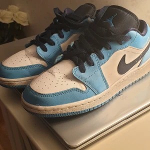  Barn Nike Air Jordan 1 Low blå/vit - Snygga barn Nike Air Jordan 1 Low sneakers i blått och vitt med svarta detaljer. Klassisk siluett med perforerad tå, svart swoosh och blå Jumpman-logga på plösen. Skorna har snörning och är tillverkade i skinn och syntet. Perfekta för dig som gillar streetstyle.