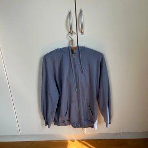 Blå hoodie med dragkedja och huva - Säljer en  blå hoodie med dragkedja framtill och stor huva. Tröjan har långa ärmar, ribbade muddar och två stora fickor på magen. Perfekt för lager-på-lager. Materialet känns som bomull och hoodien har en loose fit.