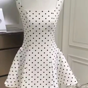 Vit prickig kortklänning med snörning - Supersöt vit kortklänning med svarta prickar och fyrkantig urringning. Klänningen har en figurnära passform upptill, utställd kjol och snygg snörning i ryggen som ger en extra detalj. Perfekt för dig som gillar retrovibbar och lekfull stil.