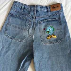 Baggy jeans från Butter - Baggy ljusblå jeans från Butter med broderade detaljer, bland annat en sko på myntfickan och en jordglob med hjärta på bakfickan. Klassisk femficksmodell i 100% bomull med raka, vida ben och hög midja. Perfekta för en avslappnad streetwear-look.