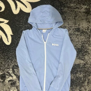 Ljusblå hoodiejacka från BOSS - Snygg ljusblå hoodiejacka från BOSS med vit dragkedja och logga på bröstet. Jackan har huva och är tillverkad i mjuk bomull, perfekt för en chill och avslappnad stil. Passar dig som gillar enkel men stilren design.