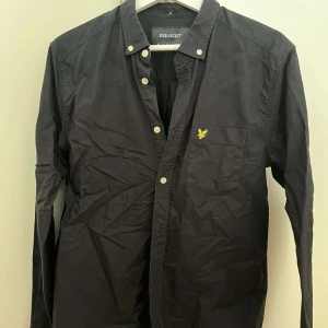 Svart skjorta från Lyle & Scott - Svart skjorta från Lyle & Scott med klassisk krage och vita knappar. Bröstficka med gul broderad logga. Långärmad och tillverkad i mjuk bomull. Perfekt för en stilren och enkel look.