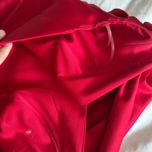 Röd axelbandslös långklänning satin - Snygg röd axelbandslös långklänning i glansigt satinliknande material. Klänningen har draperad överdel och markerad midja, med vidd i kjolen och fina veckdetaljer framtill. Perfekt för bal eller festligare tillfällen.