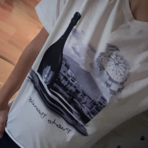 Vit t-shirt med tryck  - Vit t-shirt med coolt svartvitt tryck av en flaska, klocka och stadsmotiv på framsidan. Avslappnad passform och klassisk rund halsringning. Perfekt för dig som gillar unika prints och streetstyle. Använd 1 gång som ny 