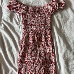 Blommig kortklänning från Chiquelle - Supersöt kortklänning från Chiquelle med blommigt mönster i rött, rosa och vitt. Klänningen har smockad överdel, volang nertill och korta ärmar. Perfekt för sommarens alla tillfällen och ger en fräsch och ungdomlig vibe.