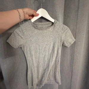 Grå basic kortärmad topp - En enkel och stilren grå topp med rund hals och korta ärmar. Modellen är figurnära och passar perfekt till jeans eller kjol. Mjuk och skön i materialet, perfekt för en clean och avslappnad look.