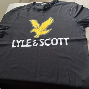 Svart Lyle & Scott t-shirt med tryck - Svart t-shirt från Lyle & Scott med stort gult örntryck och vit logga på bröstet. Klassisk rund halsringning och korta ärmar. Tillverkad i mjuk bomull, perfekt för en clean och sportig look. Nästan helt oanvänd, storlek Large.