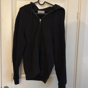 Svart hoodie med dragkedja från Zara - Svart hoodie från Zara Origins i storlek S. Klassisk modell med huva, dragsko och hel dragkedja framtill. Mjukt material och ribbade muddar vid ärmslut och nederkant. Perfekt för en chill och avslappnad stil.